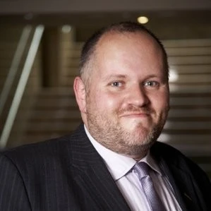 A smiling man with a beard and short hair, wearing a suit and tie, with a background of stairs.