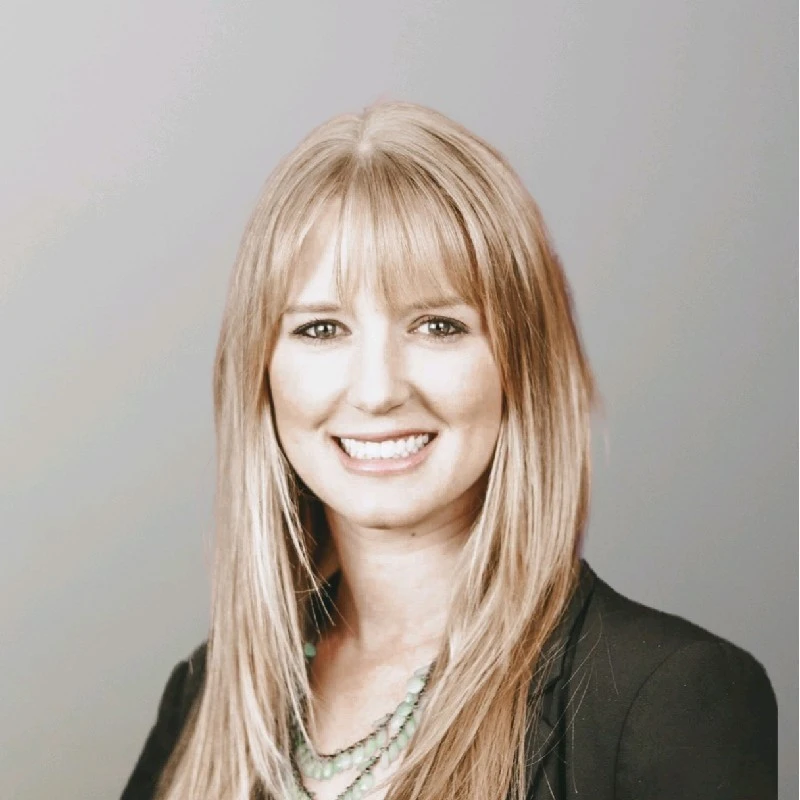 A smiling woman with long blonde hair, wearing a black blazer and necklace, against a neutral background.